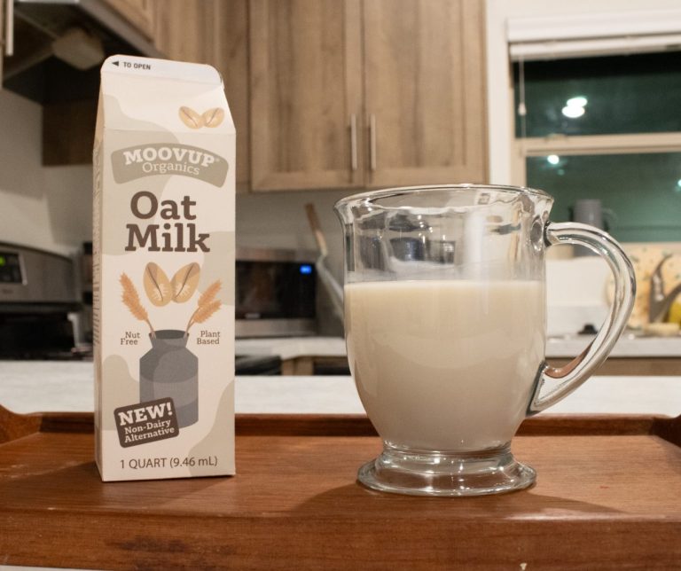 on the left is a carton of oat milk with a cow print design sitting on a tray next to a glass of oat milk in the setting of a kitchen